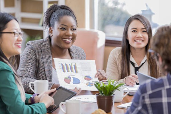 Cheerful businesswomen give presentation to male client. An African American woman is holding a chart containing bar charts and pie graphs. An Asian woman is holding a digital tablet. The client is seen from the back.